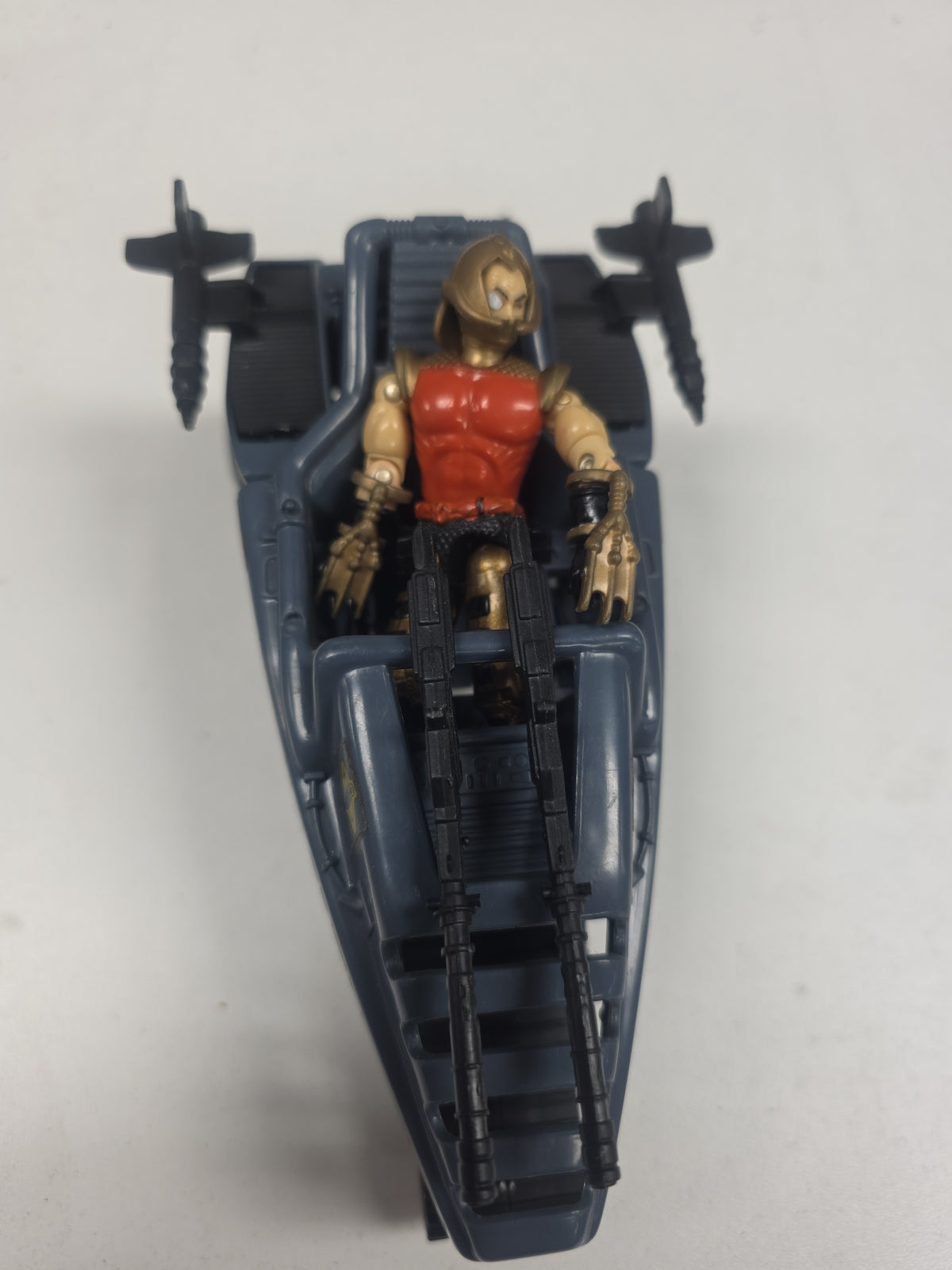 A toy action figure seated in a dark blue-gray vehicle or pod, with black weapon-like accessories and a gold-and-red figure, photographed against a plain light background.