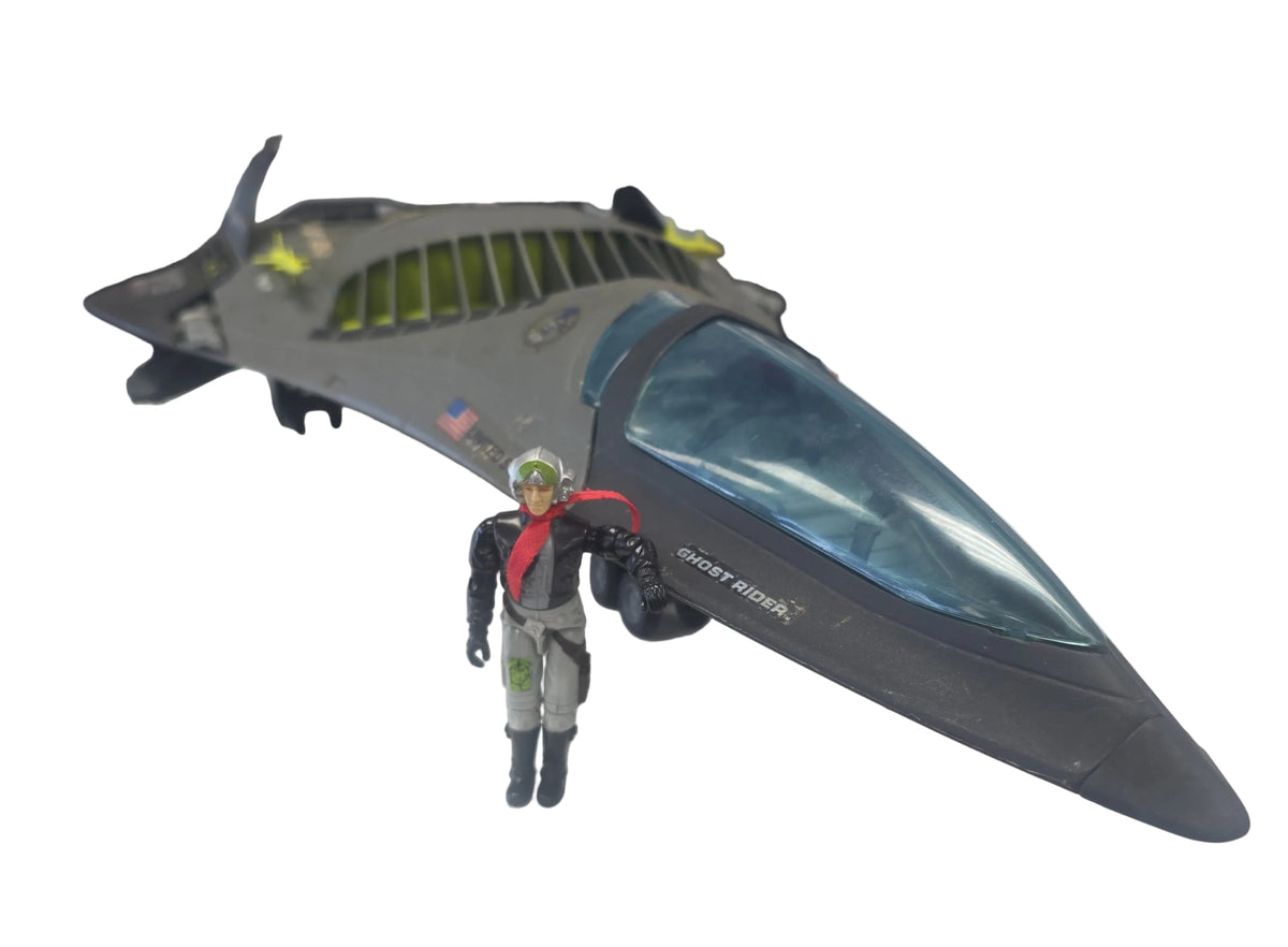 A toy model of a futuristic gray fighter jet with a blue-tinted cockpit, displayed on a white surface with a small action figure in front, against a background of comic book shelves.